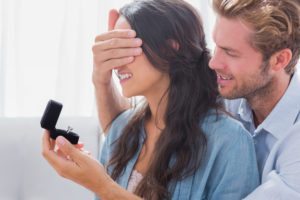 Man covers the woman's eyes from her back while holding a ring box with a small engagement ring