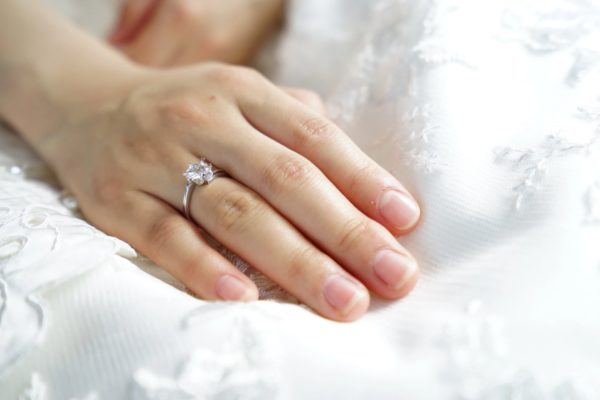 A woman's hand set against a while embroidered clothe wearing a bypass diamond engagement ring on her ring finger.