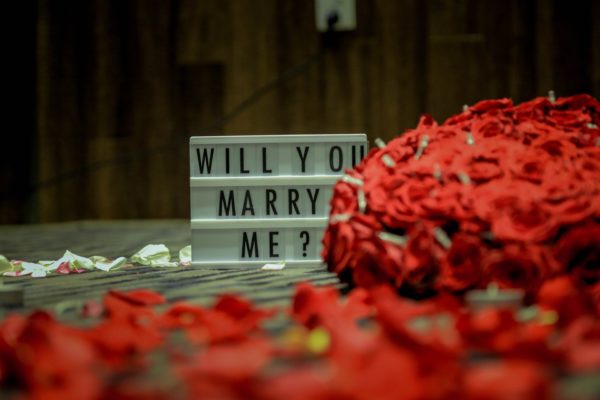 Marriage proposal ideas of selective focus photography of "Will you marry me?" board on a brown surface with a red bouquet beside