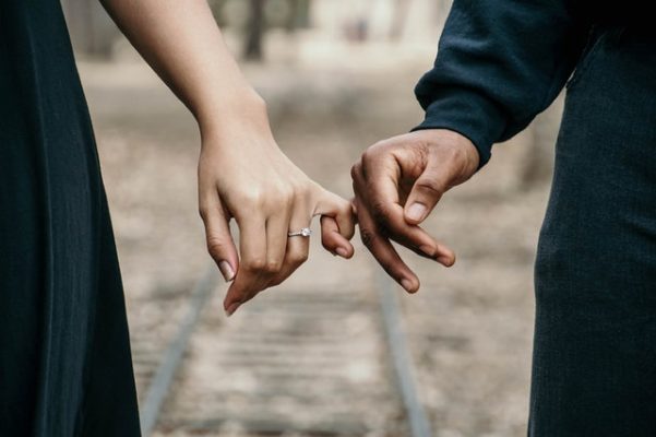 A couple standing on a railtrack with intertwined pinkies with the woman wearing her engagement ring