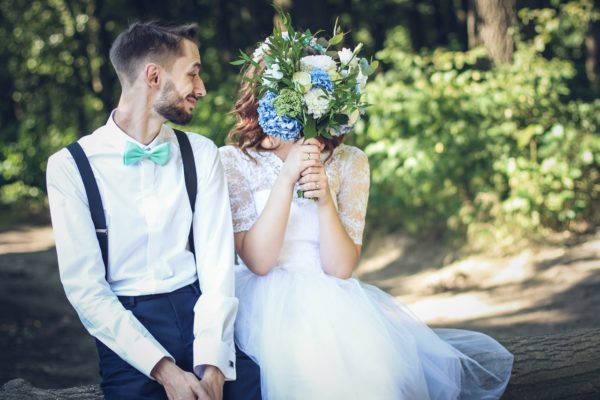 A newly wed couple sitting on a log with the girl holding a bouquet covering her face