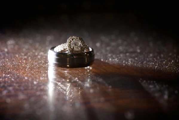 A vintage halo engageement ring placed inside a big men's wedding band, on top of a table.