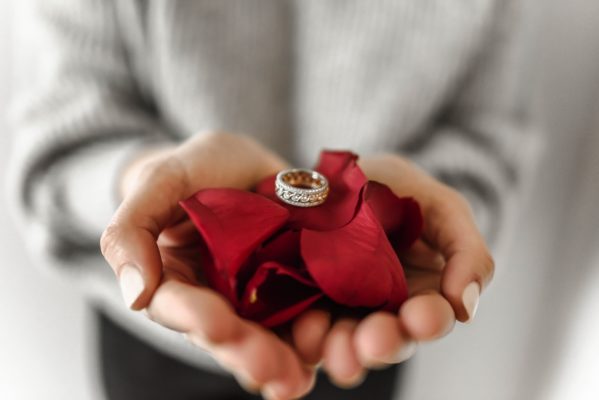 Focus view of a woman's two palm filled with red rose petals with a men's engagement ring on top