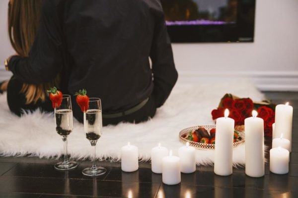 Romantic setup of a room with back view of a woman sitting in a white fluffy carpet with lit white candles and two champagne glass with strawberry