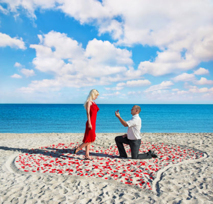 Romantic proposals of a man is kneeling in front of a woman holding a ring box beside the sea