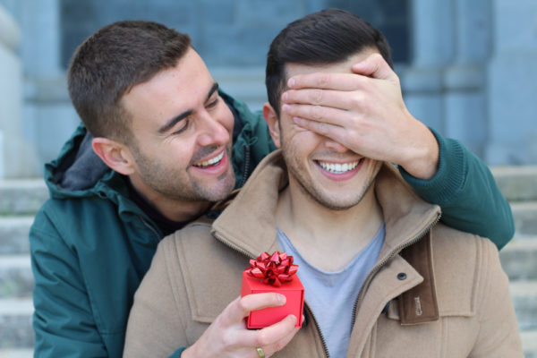 A gay proposing to his boyfriend by covering his boyfriend's eyes while holding a red engagement ring box