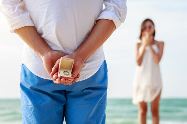 A man holding a ring box with a ring standing in front of a girl on beach