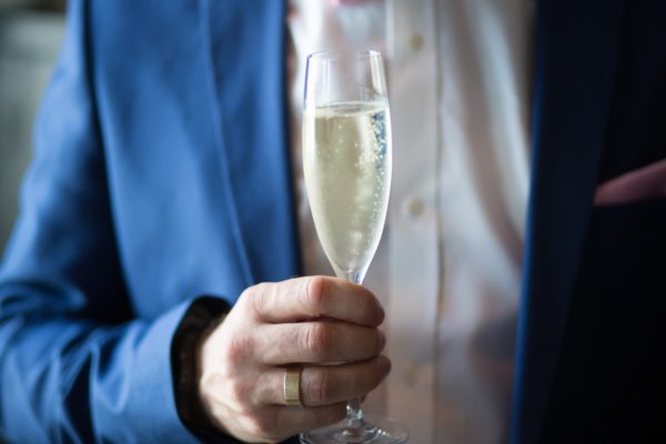 Hand of a man wearing blue suit and type of men's ring in a gold wedding band while holding a glass filled with champagne