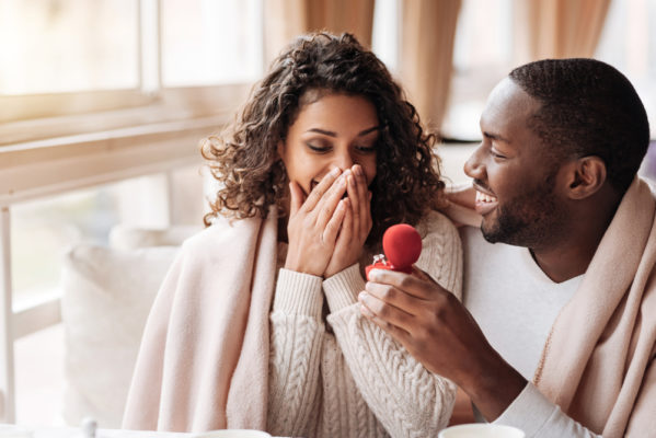 Man proposing to his fiance holding a red box with a ring imagining the engagement ring cost