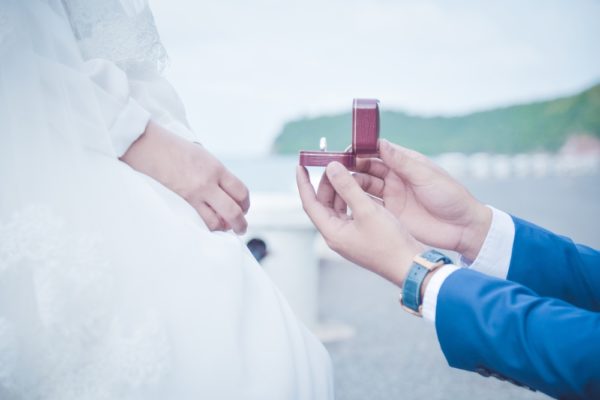 woman wearing white gown is in front of a man's hands holding a ring box with ring inside
