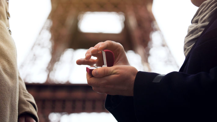 Man holding red ring box with a sparkling diamond ring kneeling in front of woman close view to Eiffel tower