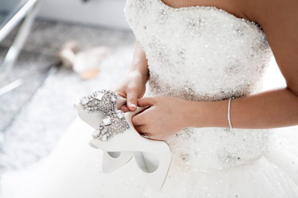 Bride wearing white gown and simple bridal jewellery holding her bridal shoes, at the back of the shoes have gemstones