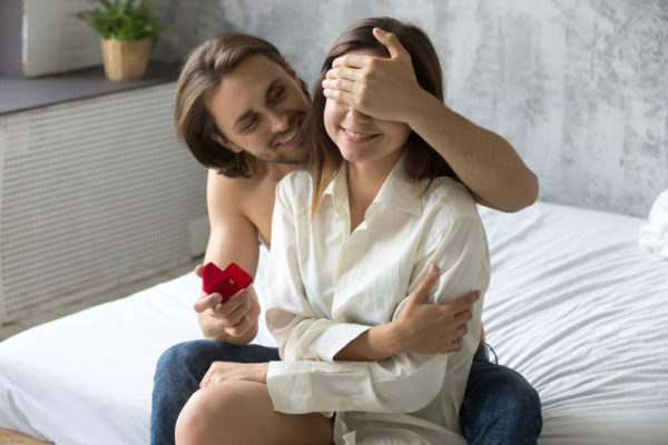 A topless man holding an engagement ring red box, covering the eyes of a woman wearing a white long sleeves