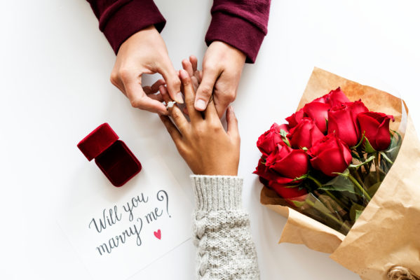 A hand of a man wears an engagement ring to a woman's ring finger, beside them are a bouquet of red roses and red box and a paper with text "will you marry me"