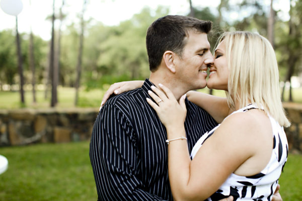 A man wearing blue striped polo kissing a woman wearing white dress and solitaire diamond ring design with bracelet
