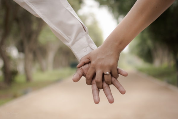 A woman's hand wearing a classic engagement rings while holding a man hand